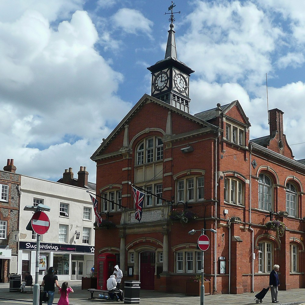 Thame Town Hall in Oxfordshire, a historic red-brick building with a clock tower, Union Jack flags, and people walking in the foreground.